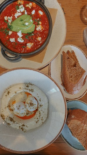 A wooden table displays a rustic meal featuring a bowl of red tomato-based stew topped with sliced avocado, crumbled cheese, and herbs. Beside it, a plate holds slices of toasted bread. Another bowl contains a creamy white dip garnished with herbs and a drizzle of oil.