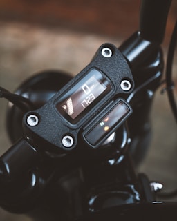 A close-up view of a motorcycle's digital dashboard displaying speed and time. The design features a modern digital interface set against a matte black finish, adorned with metal bolts.