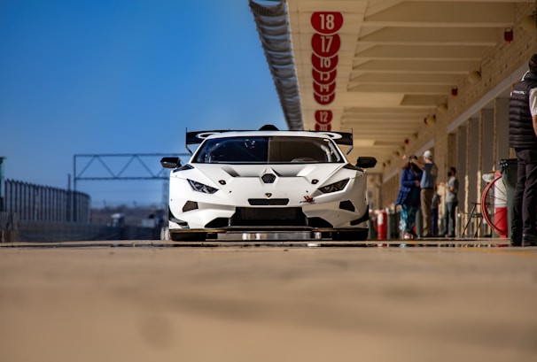 A sleek, white sports car is parked in a pit lane of a racing track. The car features aerodynamic designs and a large rear wing. Several people are visible in the background, standing next to the track infrastructure lined with numbered signs.