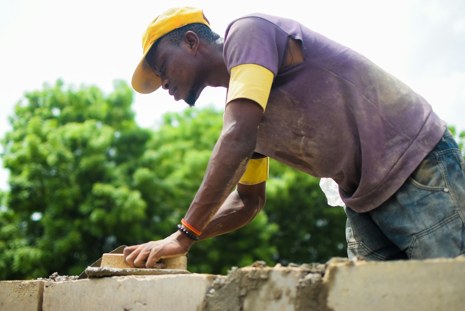 A mason in a ball cap smoothing mortar along the top of a block wall on a sunny summer jobsite.