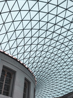 A geometric, glass-paneled roof forms a dramatic grid of triangles creating an intricate pattern. The structure is part of a large, curved building wall with classical architectural elements such as arches and columns. Natural light filters through the roof, casting subtle reflections.