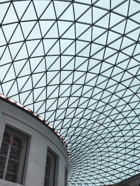 A geometric, glass-paneled roof forms a dramatic grid of triangles creating an intricate pattern. The structure is part of a large, curved building wall with classical architectural elements such as arches and columns. Natural light filters through the roof, casting subtle reflections.