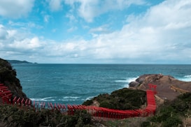 A coastal landscape with a bright blue sky scattered with clouds. A series of red torii gates form a path that winds along the rocky shoreline, surrounded by lush green vegetation. The ocean stretches out to the horizon with gentle waves creating a serene atmosphere.