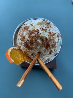 A creamy dessert garnished with whipped cream, a sprinkle of chocolate flakes, a slice of orange, and two pretzel sticks, served in a cup placed on a wooden coaster.