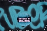 A circular graphic overlays a graffiti background with the phrase 'FUTURE IS CREATIVE' displayed in bold, neon pink letters on a cracked, textured blue surface.
