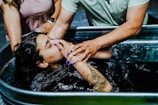 A person with tattoos is being baptized in a large metal container filled with water. A hand is supporting their head and covering their face to help with the immersion. The scene involves multiple individuals participating in the ceremony, one of whom has colorful nails.