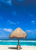 A tranquil beach scene featuring a thatched umbrella on the sandy shore. The turquoise ocean waves gently break in the background under a sky dotted with a few fluffy clouds. A person is sitting underneath the umbrella, partially obscured.