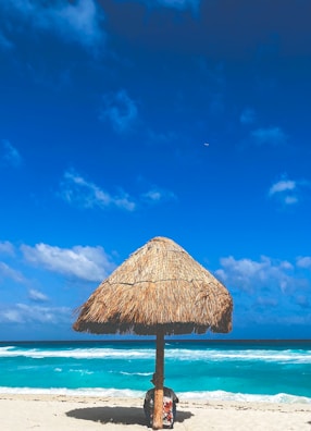 A tranquil beach scene featuring a thatched umbrella on the sandy shore. The turquoise ocean waves gently break in the background under a sky dotted with a few fluffy clouds. A person is sitting underneath the umbrella, partially obscured.