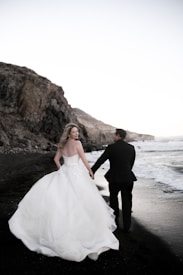 A couple is walking hand in hand along a rocky beach. The woman is wearing a flowing white wedding dress while the man is in a dark suit. The sea stretches out beside them, with waves gently lapping at the shore. Tall cliffs rise along the coastline, creating a dramatic backdrop.