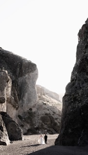 A couple dressed in formal attire walks hand in hand through a rugged landscape surrounded by towering rock formations. The lighting is soft, diffused by the bright sky above, casting gentle shadows on the sandy ground.