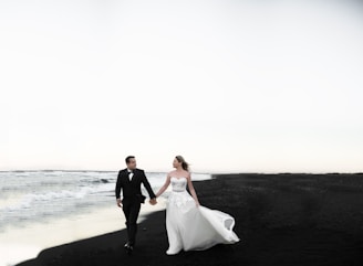 A bride and groom walk hand in hand along a deserted beach with dark sand. The bride wears a flowing white gown, while the groom is dressed in a black suit. Gentle waves roll onto the shore, and the sky above is overcast, contributing to a serene and slightly dramatic setting.