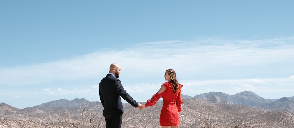 man in black suit jacket holding woman in red dress on top of mountain during daytime