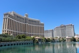 A large luxury hotel and casino building with multiple stories, featuring a light facade with arched windows. The structure has a prominent central tower with a dome-like top. It is surrounded by landscaping with trees and a body of water in the foreground.