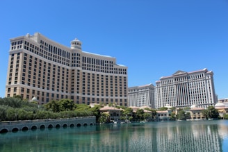 A large luxury hotel and casino building with multiple stories, featuring a light facade with arched windows. The structure has a prominent central tower with a dome-like top. It is surrounded by landscaping with trees and a body of water in the foreground.