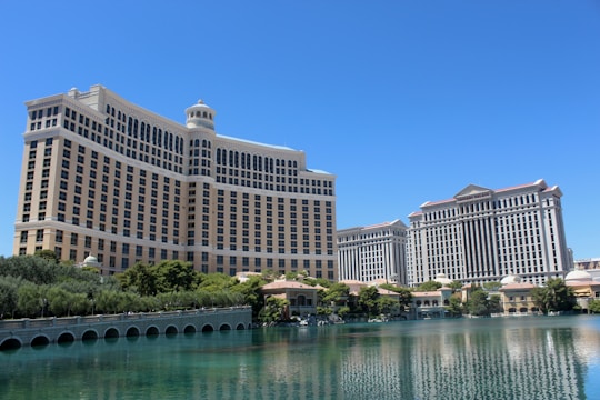 A large luxury hotel and casino building with multiple stories, featuring a light facade with arched windows. The structure has a prominent central tower with a dome-like top. It is surrounded by landscaping with trees and a body of water in the foreground.
