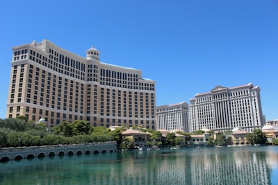 A large luxury hotel and casino building with multiple stories, featuring a light facade with arched windows. The structure has a prominent central tower with a dome-like top. It is surrounded by landscaping with trees and a body of water in the foreground.