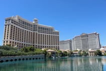A large luxury hotel and casino building with multiple stories, featuring a light facade with arched windows. The structure has a prominent central tower with a dome-like top. It is surrounded by landscaping with trees and a body of water in the foreground.