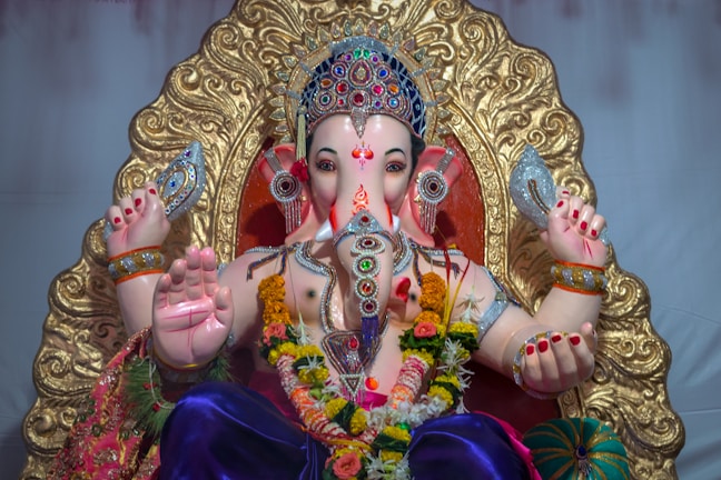 A detailed statue of a deity adorned with ornate jewelry and garments. The figure is richly decorated with vibrant flowers and intricate patterns, sitting against a backdrop with a golden, ornate design. The hands hold various symbolic objects, and the face is intricately painted with a serene expression.