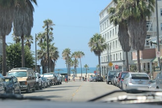 A sunny coastal street lined with palm trees and parked cars leads towards the beach. The street is flanked by multi-story buildings, and a few people can be seen heading towards the ocean. The blue sky and ocean create a bright and inviting atmosphere.