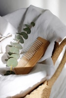 A wooden comb rests on a soft, white cloth inside a woven basket. A sprig of eucalyptus lies next to the comb, adding a touch of greenery.