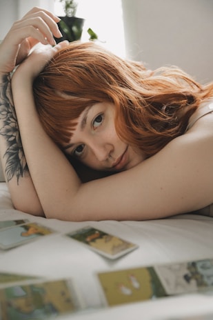 A person with red hair rests their head on their arm while lying down. They have a tattoo on their arm and appear relaxed. Several tarot cards are spread out on the surface nearby.