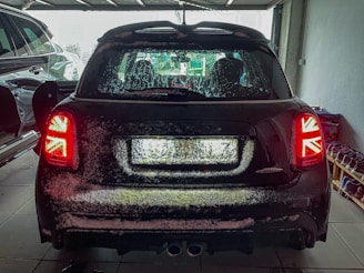 A car with a snow-covered rear is parked inside a garage. The tail lights feature a Union Jack design. The garage is dimly lit, with another car partially visible on the left and a rack of shoes on the right.