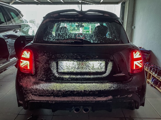 A car with a snow-covered rear is parked inside a garage. The tail lights feature a Union Jack design. The garage is dimly lit, with another car partially visible on the left and a rack of shoes on the right.