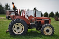 A vintage orange Zetor tractor is parked on a green grassy field. The tractor has large black tires and is positioned sideways, revealing the worn details on its body. In the background, there's an old truck and shipping containers, along with some trees.