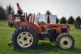 A vintage orange Zetor tractor is parked on a green grassy field. The tractor has large black tires and is positioned sideways, revealing the worn details on its body. In the background, there's an old truck and shipping containers, along with some trees.