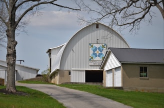 A large white barn with a curved roof and a quilt square design featuring blue flowers and green leaves on the front, commemorating Clinton County's bicentennial. It sits on a slight grassy hill, with a winding driveway leading up to it, bordered by bare trees. Adjacent to the barn is a smaller attached structure with a dark roof and garage door. The sky is overcast, giving the scene a calm and muted appearance.