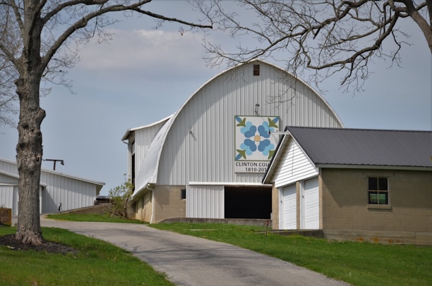 A large white barn with a curved roof and a quilt square design featuring blue flowers and green leaves on the front, commemorating Clinton County's bicentennial. It sits on a slight grassy hill, with a winding driveway leading up to it, bordered by bare trees. Adjacent to the barn is a smaller attached structure with a dark roof and garage door. The sky is overcast, giving the scene a calm and muted appearance.