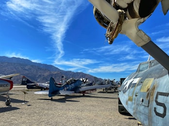 A collection of vintage military aircraft is parked on an open-air tarmac under a bright blue sky with wispy clouds. The aircraft, displaying various markings and numbers, are set against a backdrop of rugged mountains.