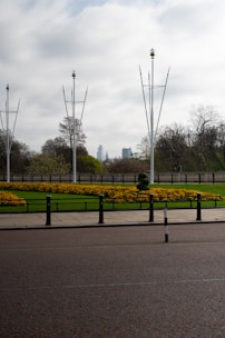 A landscaped garden with neatly arranged yellow flowers on lush green grass, bordered by a stone fence. Tall, slender metal poles with decorative elements rise into a cloudy sky. In the distance, skyscrapers can be seen, partially obscured by trees. The foreground features a paved road with black and white bollards.