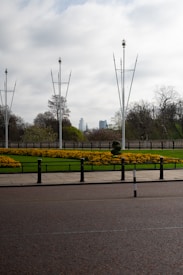 A landscaped garden with neatly arranged yellow flowers on lush green grass, bordered by a stone fence. Tall, slender metal poles with decorative elements rise into a cloudy sky. In the distance, skyscrapers can be seen, partially obscured by trees. The foreground features a paved road with black and white bollards.