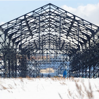 A large, industrial metal framework structure is set against a snowy landscape. The intricate lattice of steel beams forms a symmetrical pattern, with a distant cityscape visible through the framework. The ground is blanketed in snow, dotted with dry grass.