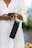 A happy young woman holding her black stainless steel water bottle with a smartphone attached, outdoors.