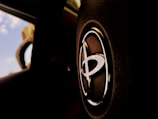 A close-up view of a car's steering wheel displaying a prominent Toyota logo in a well-lit interior. The background is blurred, showing part of the windshield and a glimpse of the blue sky.