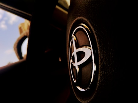A close-up view of a car's steering wheel displaying a prominent Toyota logo in a well-lit interior. The background is blurred, showing part of the windshield and a glimpse of the blue sky.