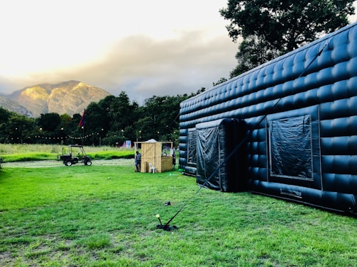 A large inflatable black structure is set up on a green grassy field. In the background, there is a small tractor and a wooden stall with people gathered around it. Tall trees border the area, with mountain peaks visible in the distance under a cloudy sky.