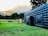 A large inflatable black structure is set up on a green grassy field. In the background, there is a small tractor and a wooden stall with people gathered around it. Tall trees border the area, with mountain peaks visible in the distance under a cloudy sky.