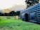 A large inflatable black structure is set up on a green grassy field. In the background, there is a small tractor and a wooden stall with people gathered around it. Tall trees border the area, with mountain peaks visible in the distance under a cloudy sky.