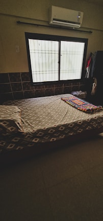 A bedroom with a single bed covered by a patterned bedspread. There is a folded, colorful bed mat on the bed. The room has a window with bars, partially covered by a sheer curtain. An air conditioning unit is installed above the window. Dark tiles can be seen on the lower part of the wall, and clothes hang to the right of the window.