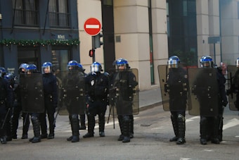 A group of uniformed riot police officers stand in formation on a street near an intersection. Each officer is equipped with a transparent riot shield, a helmet with a visor, and protective gear including knee pads. A 'no entry' traffic sign is visible in the background alongside a building adorned with greenery on the exterior.