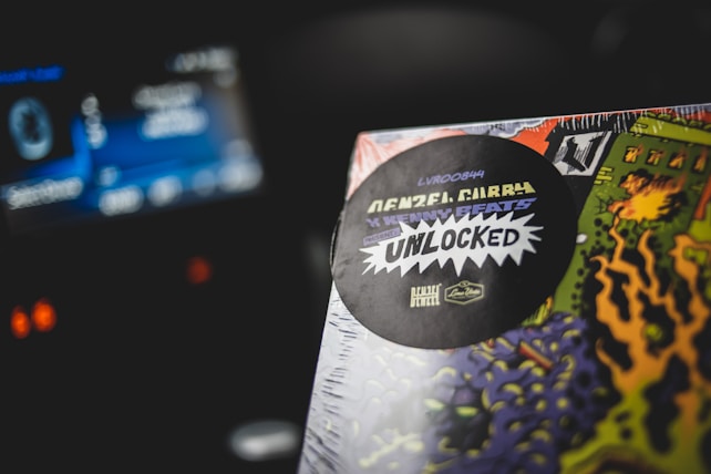 A close-up of a colorful album cover with a black sticker displaying text. The text includes 'Denzel Curry' and 'Unlocked' in prominent font. The background features vibrant artistic designs in green, orange, and purple hues. The setting appears to be inside a car with a blurred dashboard view.