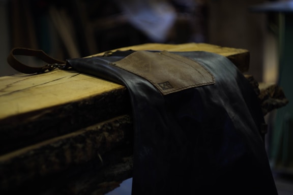 A leather apron is draped over a varnished wooden slab with a textured edge. The scene is dimly lit, emphasizing the rustic and craft-focused atmosphere of the setting.