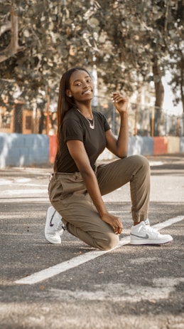 A person kneeling on one knee on an outdoor basketball court, surrounded by trees with blurred foliage. Wearing a black t-shirt, light brown pants, and white sneakers. Sunlight creates a warm, serene atmosphere.