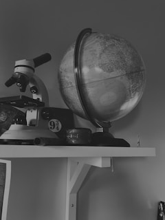 A black and white image featuring a globe on a shelf alongside a microscope. Below the microscope, there is a small circular sign displaying the number 9 3/4. The setting appears to be a study or an academic environment. Various other objects are visible but not easily identifiable due to the limited color palette.