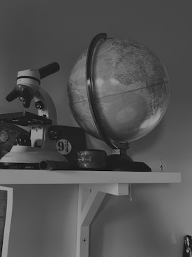 A black and white image featuring a globe on a shelf alongside a microscope. Below the microscope, there is a small circular sign displaying the number 9 3/4. The setting appears to be a study or an academic environment. Various other objects are visible but not easily identifiable due to the limited color palette.