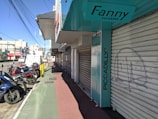 A city street view featuring several parked motorcycles and scooters near closed storefronts. The store signs include 'Fanny Calçados' and 'Piccadilly'. The sidewalk is divided into sections with green and red paving. Overhead, numerous power lines stretch across the sky.