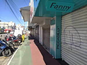 A city street view featuring several parked motorcycles and scooters near closed storefronts. The store signs include 'Fanny Calçados' and 'Piccadilly'. The sidewalk is divided into sections with green and red paving. Overhead, numerous power lines stretch across the sky.